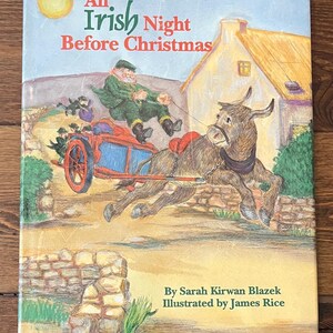 Op de afbeelding: Kinderboek getiteld "An Irish Night Before Christmas" met een illustratie van een man in groene kleding die in een kar rijdt die door een ezel wordt getrokken. De cover toont een gele zon en een huisje op de achtergrond.