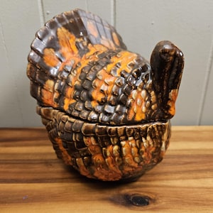 May include: A ceramic turkey-shaped container with a lid. The turkey is primarily brown, orange, and black, with a textured, scale-like pattern. The container is likely used for decorative or storage purposes.