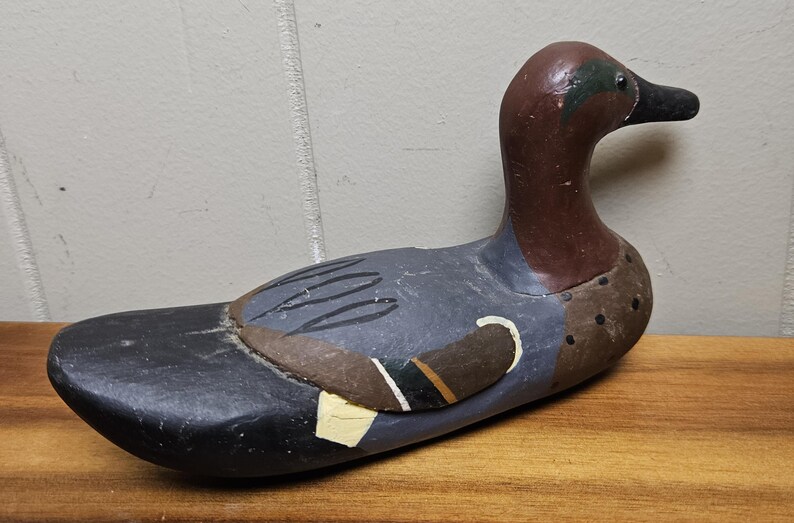 May include: A hand-painted wooden duck decoy. The duck has a brown head and neck, a gray body with black markings, and a black tail. The decoy is resting on a wooden surface.