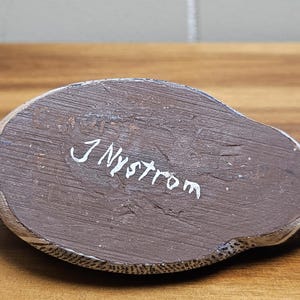 Op de afbeelding: Een handgesneden houten eendenbeeldje, van onderaf gezien. Het lichaam van de eend is bruin met witte en zwarte accenten. De handtekening van de kunstenaar, "J Nystrom", staat in witte letters op de basis. De eend heeft een kleine, zwarte staart.