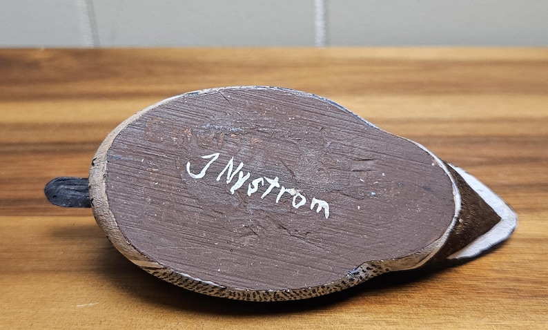 Op de afbeelding: Een handgesneden houten eendenbeeldje, van onderaf gezien. Het lichaam van de eend is bruin met witte en zwarte accenten. De handtekening van de kunstenaar, "J Nystrom", staat in witte letters op de basis. De eend heeft een kleine, zwarte staart.