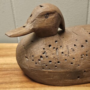 May include: A detailed, hand-carved wooden duck decoy. The duck is brown with visible wood grain and numerous small holes. The beak is long and pointed, and the eye is a small metal piece. The decoy sits on a wooden surface.