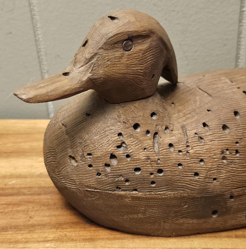 May include: A detailed, hand-carved wooden duck decoy. The duck is brown with visible wood grain and numerous small holes. The beak is long and pointed, and the eye is a small metal piece. The decoy sits on a wooden surface.