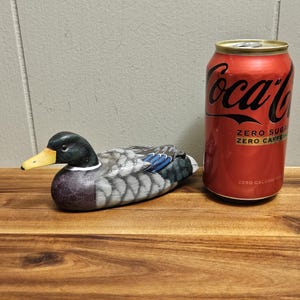 Puede incluir: Un se&ntilde;uelo de pato de madera con cabeza negra, pico amarillo y plumas grises y azules. El pato est&aacute; sobre una superficie de madera junto a una lata roja de Coca-Cola con el texto "ZERO SUGAR ZERO CAFFEINE".
