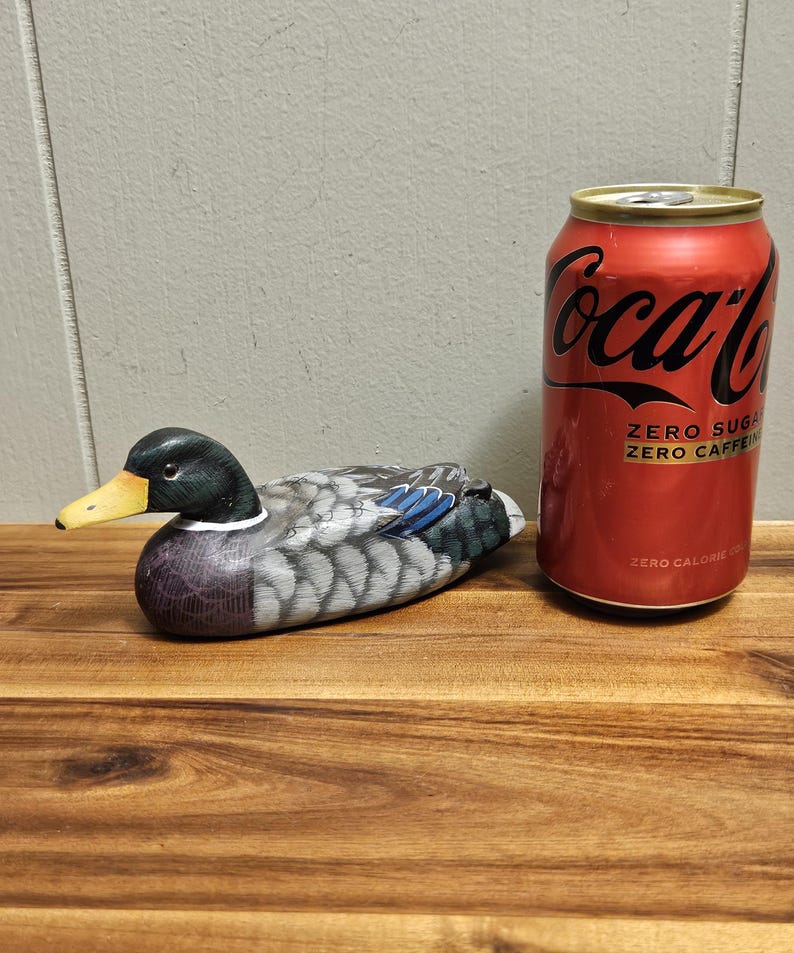 Puede incluir: Un se&ntilde;uelo de pato de madera con cabeza negra, pico amarillo y plumas grises y azules. El pato est&aacute; sobre una superficie de madera junto a una lata roja de Coca-Cola con el texto "ZERO SUGAR ZERO CAFFEINE".