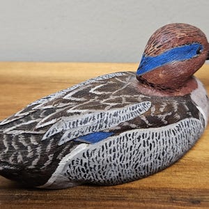 Op de afbeelding: Een handgesneden houten eendenbeeldje. De eend heeft een bruine kop met een blauwe streep, een zwarte snavel en een lichaam met grijze, witte en bruine veren. Het beeldje rust op een houten oppervlak.