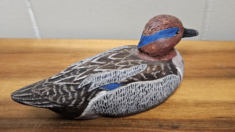 Op de afbeelding: Een handgesneden houten eendenbeeldje. De eend heeft een bruine kop met een blauwe streep, een zwarte snavel en een lichaam met grijze, witte en bruine veren. Het beeldje rust op een houten oppervlak.