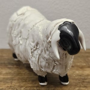 May include: A ceramic figurine of a Valais Blacknose sheep with a glossy white body featuring textured, layered wool details. The sheep has a smooth, solid black face and ears, with curved white horns extending from the sides of its head.
