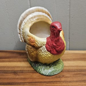 Puede incluir: Una maceta de cerámica con forma de pavo, con cabeza y carúnculas rojas, cuerpo amarillo y naranja, y cola en abanico blanca y marrón. La maceta se asienta sobre una base verde, que se asemeja a hierba. Tiene una cavidad abierta.