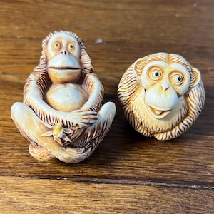 May include: Two monkey figurines. One is seated with arms crossed, holding a small yellow object. The other is a round monkey head. Both are tan and brown, with detailed facial features and fur-like textures.