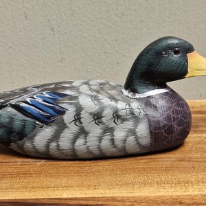 Puede incluir: Figura de pato de madera pintada a mano. El pato tiene una cabeza negra, un pico amarillo y un cuerpo con detalles grises, azules y morados. La figura descansa sobre una superficie de madera.
