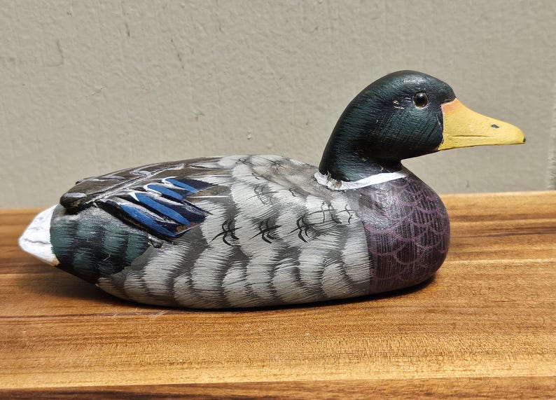 Puede incluir: Figura de pato de madera pintada a mano. El pato tiene una cabeza negra, un pico amarillo y un cuerpo con detalles grises, azules y morados. La figura descansa sobre una superficie de madera.