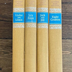 May include: Four vintage books by Louisa May Alcott. The spines are mustard yellow with blue title blocks. Titles include "Under the Lilacs", "Jo's Boys", "Jack and Jill", and "Eight Cousins". The publisher is Nelson Doubleday.