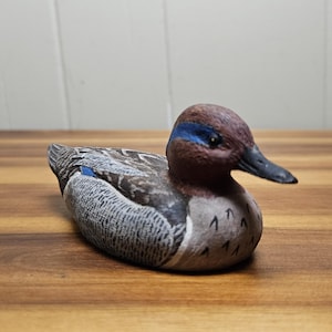Op de afbeelding: Houten eendenbeeldje met gedetailleerde veermotieven in tinten grijs, bruin en blauw. De eend heeft een bruine kop met een blauwe streep en een zwarte snavel. Het beeldje ligt op een houten oppervlak.