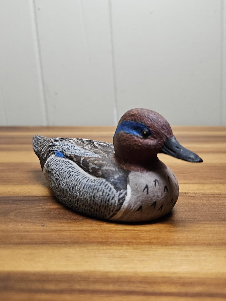 Op de afbeelding: Houten eendenbeeldje met gedetailleerde veermotieven in tinten grijs, bruin en blauw. De eend heeft een bruine kop met een blauwe streep en een zwarte snavel. Het beeldje ligt op een houten oppervlak.