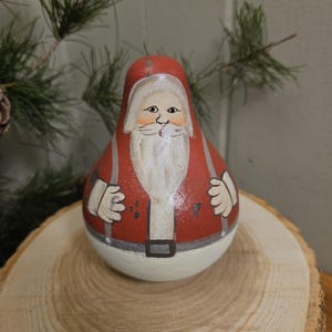 May include: A hand-painted Santa Claus figurine with a red coat, white beard, and a white base. The Santa Claus is standing on a wooden slice, with pine branches in the background. The figurine has a rustic, folk art style.