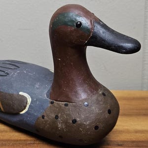 May include: A hand-painted wooden duck decoy. The duck has a brown head with a green stripe, a black beak, and a blue-gray body. The decoy has black dots on the chest and is resting on a wooden surface.