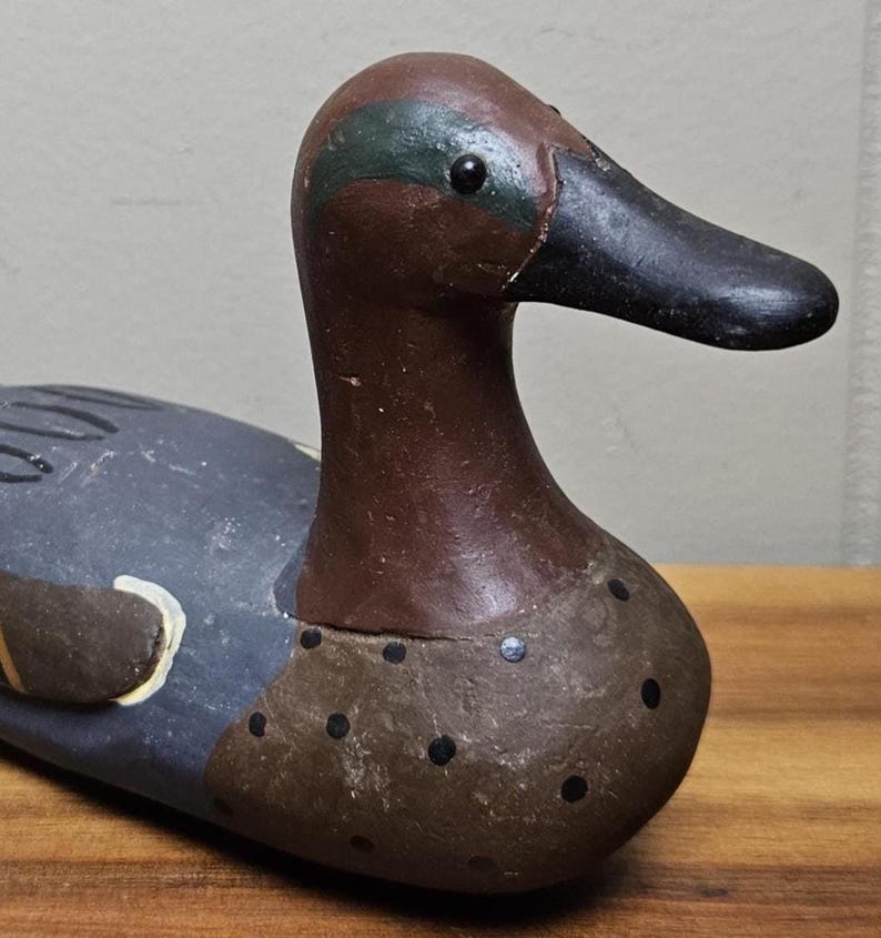 May include: A hand-painted wooden duck decoy. The duck has a brown head with a green stripe, a black beak, and a blue-gray body. The decoy has black dots on the chest and is resting on a wooden surface.