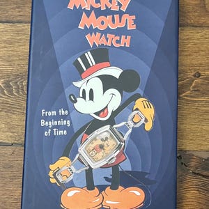 May include: A navy blue book cover featuring Mickey Mouse holding a watch. The title "The Mickey Mouse Watch" is in orange and white text. The book cover also includes the text "From the Beginning of Time" and the names Robert Heide & John Gilman.