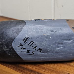 May include: A hand-painted wooden duck decoy. The decoy is painted in shades of black, gray, and brown, with black dots on the brown section. The name "William Tess" is handwritten on the side. The decoy is resting on a wooden surface.