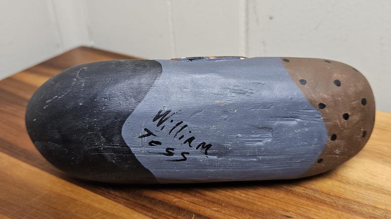May include: A hand-painted wooden duck decoy. The decoy is painted in shades of black, gray, and brown, with black dots on the brown section. The name "William Tess" is handwritten on the side. The decoy is resting on a wooden surface.