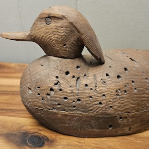 May include: A vintage wooden duck decoy, hand-carved with visible wood grain and numerous small holes. The duck has a detailed head and beak, and is painted in shades of brown. This rustic piece is a classic example of folk art.