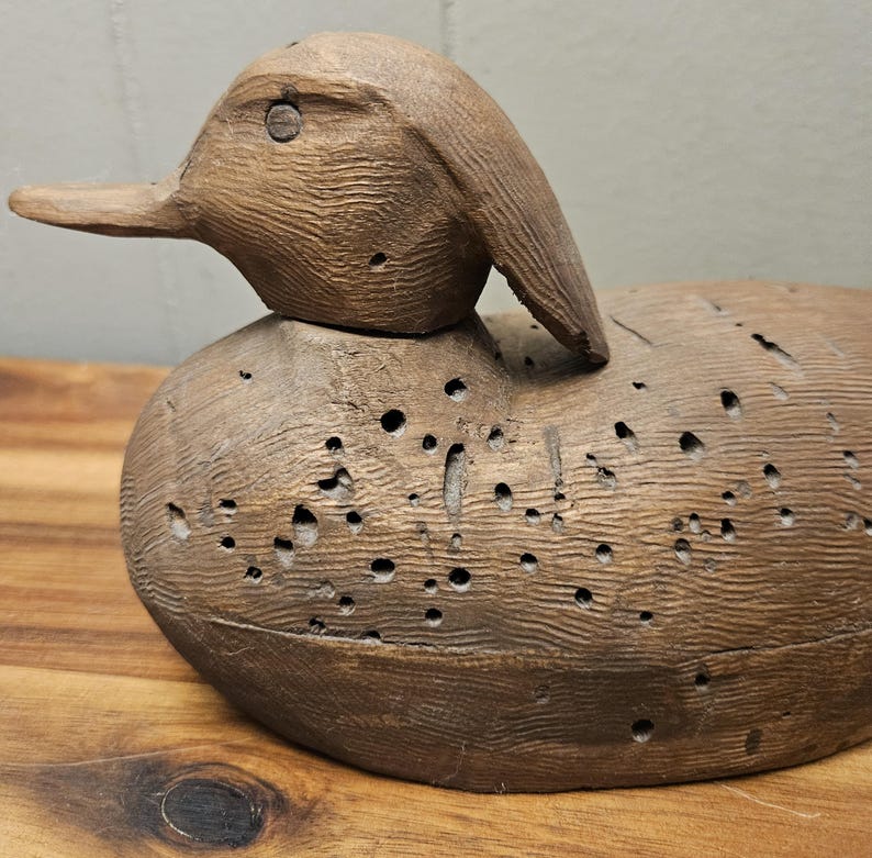 May include: A vintage wooden duck decoy, hand-carved with visible wood grain and numerous small holes. The duck has a detailed head and beak, and is painted in shades of brown. This rustic piece is a classic example of folk art.