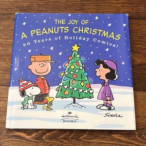 May include: A Peanuts Christmas book cover with a blue background and white stars. The cover features Charlie Brown, Snoopy, Lucy, and a decorated Christmas tree. Text reads "The Joy of A Peanuts Christmas" and "50 Years of Holiday Comics!"