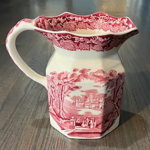 May include: Cream-colored pitcher with a red scenic design. The pitcher features a handle and a scalloped rim with a floral pattern. The main body of the pitcher displays a red and white landscape scene with trees, a castle, and figures.