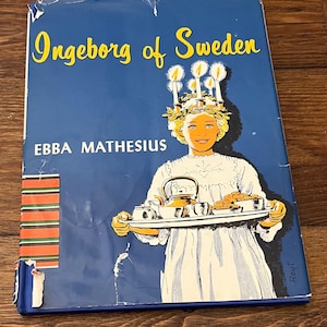 以下が含まれることがあります： 青い背景に黄色の文字で「Ingeborg of Sweden」と書かれたヴィンテージ本の表紙。表紙には、ろうそくの王冠をかぶり、ティーセットとペストリーが載ったトレイを持つ少女のイラストが描かれています。