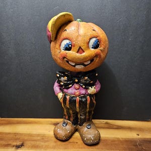 May include: A decorative figurine featuring a pumpkin-headed character wearing a yellow cap, a black bow tie with white dots, a purple shirt with orange buttons, and black and orange striped trousers, finished with brown shoes with white speckles.