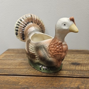 Puede incluir: Un macetero o plato de servicio de cerámica con forma de pavo que presenta una cabeza blanca, un pico amarillo y un cuerpo con plumas marrones, bronceadas y negras. El pavo tiene una cola en abanico y un cuello texturizado con una cavidad.