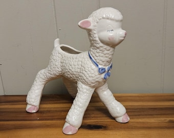 May include: A white ceramic lamb planter with a textured body, pink accents on the ears and hooves, and a blue ribbon around its neck. The lamb has a small opening on its back for planting. The planter is on a wooden surface.