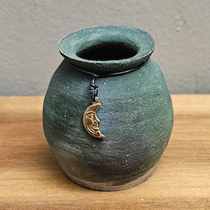 May include: A small, dark green ceramic vase with a rounded body and a wide opening. A gold-colored crescent moon charm hangs from the rim. The vase is displayed on a wooden surface.