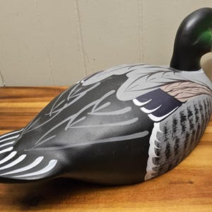 May include: A decorative duck decoy with a black head and body, featuring gray and white wing details. The duck has a white beak and is positioned on a wooden surface. The decoy is a classic piece of home decor.