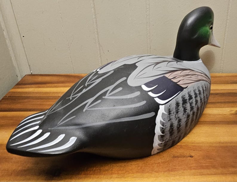 May include: A decorative duck decoy with a black head and body, featuring gray and white wing details. The duck has a white beak and is positioned on a wooden surface. The decoy is a classic piece of home decor.