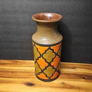 Op de afbeelding: Een cilindrische keramische vaas met een uitlopende rand, voorzien van een textuurrijke bruine hals en een lichaam versierd met een oranje en olijfgroen vierpaspatroon omlijnd in zwart. De voet van de vaas is plat en de binnenkant is glad bruin.