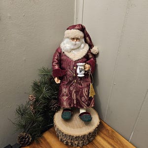 Puede incluir: Figura decorativa de Papá Noel con una túnica y sombrero burdeos con estampado de cachemira y ribete de piel color crema. La figura sostiene una taza y está sobre una rodaja de madera. Una ramita de pino y piñas están cerca.