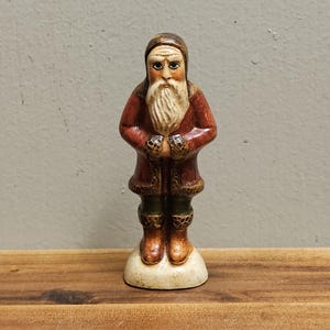 Könnte beinhalten: Eine Keramikfigur von Weihnachtsmann, der einen roten Mantel mit weißem Pelzbesatz, einen weißen Bart und braune Stiefel trägt. Er steht auf einem weißen Sockel.