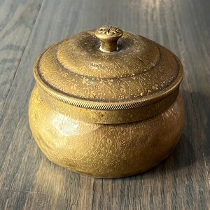 May include: A small, round, brass trinket box with a floral design and a knob on the lid.