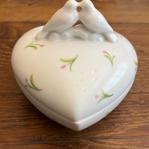 May include: A white heart-shaped trinket box with a lid featuring two kissing doves. The box is decorated with small pink flowers and green leaves. The box is made of porcelain.