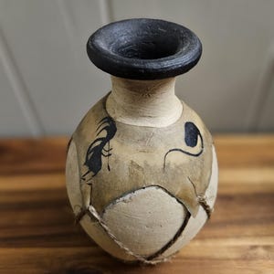 Puede incluir: Jarrón de cerámica hecho a mano con borde negro y diseños abstractos negros sobre un cuerpo de color beige y crema. El jarrón tiene una apariencia texturizada única con costuras visibles y detalles de cuerda, mostrando una estética rústica.