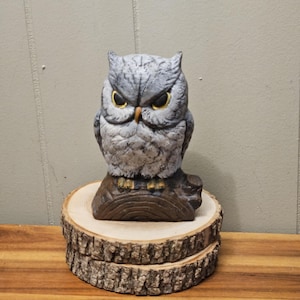 May include: A gray owl figurine with yellow eyes and a brown beak, perched on a wooden base. The owl is detailed with textured feathers and sits atop a natural wood slice, creating a rustic aesthetic.