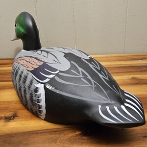 May include: A hand-painted wooden duck decoy. The duck features a green head, black neck, and a body with gray, black, and white painted details. The decoy is positioned on a wooden surface, showcasing its detailed craftsmanship.