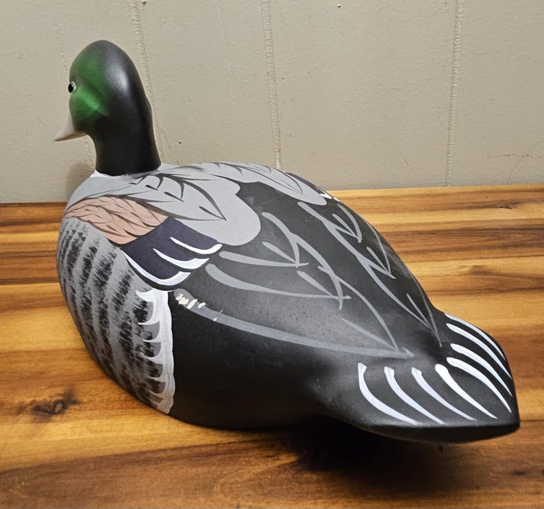 May include: A hand-painted wooden duck decoy. The duck features a green head, black neck, and a body with gray, black, and white painted details. The decoy is positioned on a wooden surface, showcasing its detailed craftsmanship.