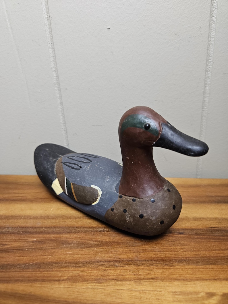 May include: A wooden duck decoy with a brown head and neck, a black beak, and a blue-gray body. The decoy has painted details, including a white and yellow wing marking. The duck is resting on a wooden surface.