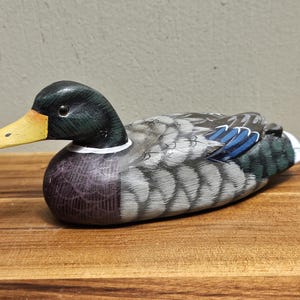 Puede incluir: Figura de pato de madera pintada a mano. El pato tiene una cabeza negra con un pico amarillo, un cuello blanco y un cuerpo con detalles grises, azules y morados. El pato est&aacute; sobre una superficie de madera.