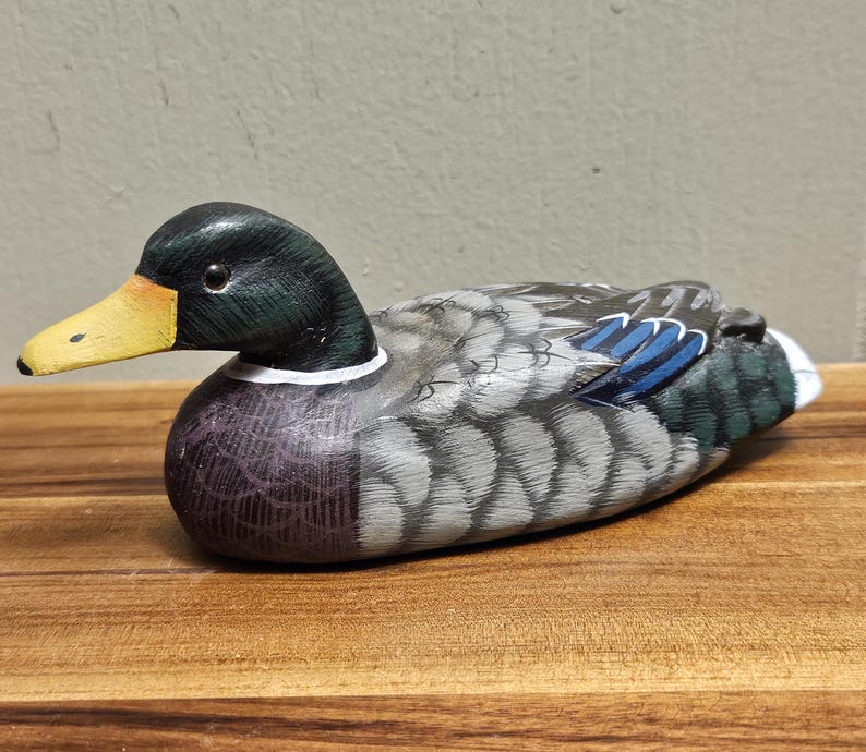 Puede incluir: Figura de pato de madera pintada a mano. El pato tiene una cabeza negra con un pico amarillo, un cuello blanco y un cuerpo con detalles grises, azules y morados. El pato est&aacute; sobre una superficie de madera.