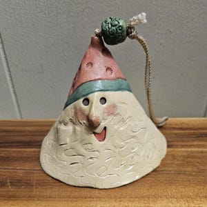 May include: A ceramic Christmas ornament shaped like a Santa Claus bell with a red hat, green trim, and a white beard. The ornament has a string for hanging and a green bead at the top.