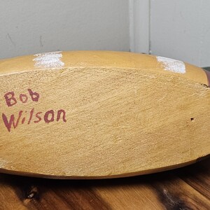 K&ouml;nnte beinhalten: H&ouml;lzerner Entenk&ouml;der mit lackiertem Finish. Die Unterseite ist sichtbar und zeigt den Text "Bob Wilson" in Rot. Die Ente hat einen beigen K&ouml;rper, wei&szlig;e und schwarze Akzente und einen braunen Schwanz.
